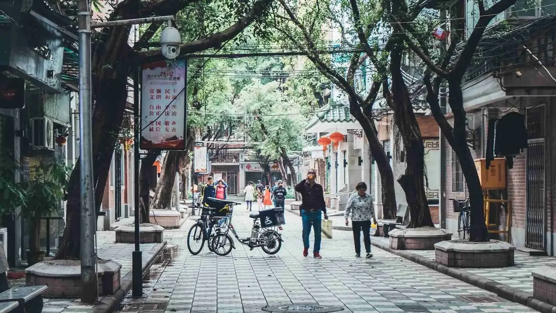 A quiet, tree-lined pedestrian street in a Chinese city, with people walking past traditional shopfronts, red lanterns and a CCTV surveillance camera mounted on a pole.