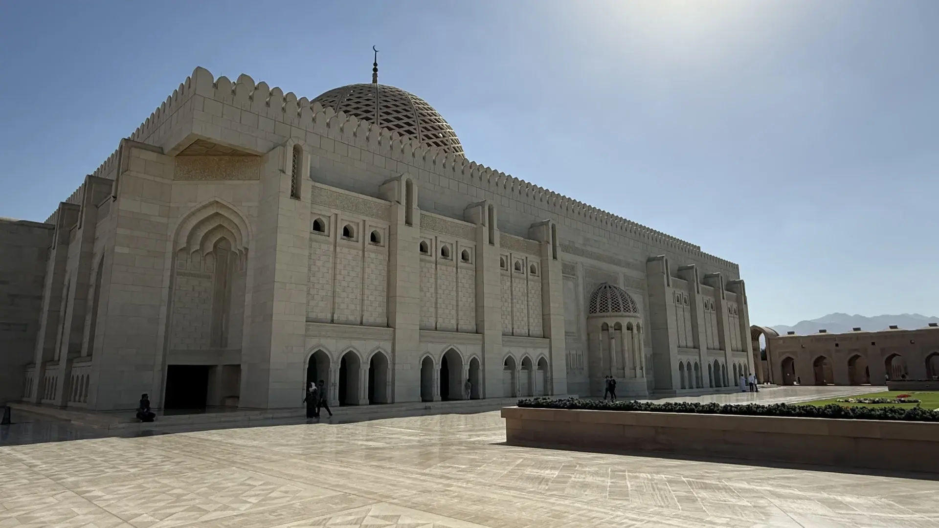 A wide-angle view of the intricate limestone architecture and central dome of the Sultan Qaboos Grand Mosque in Muscat, Oman, set against a clear blue sky.