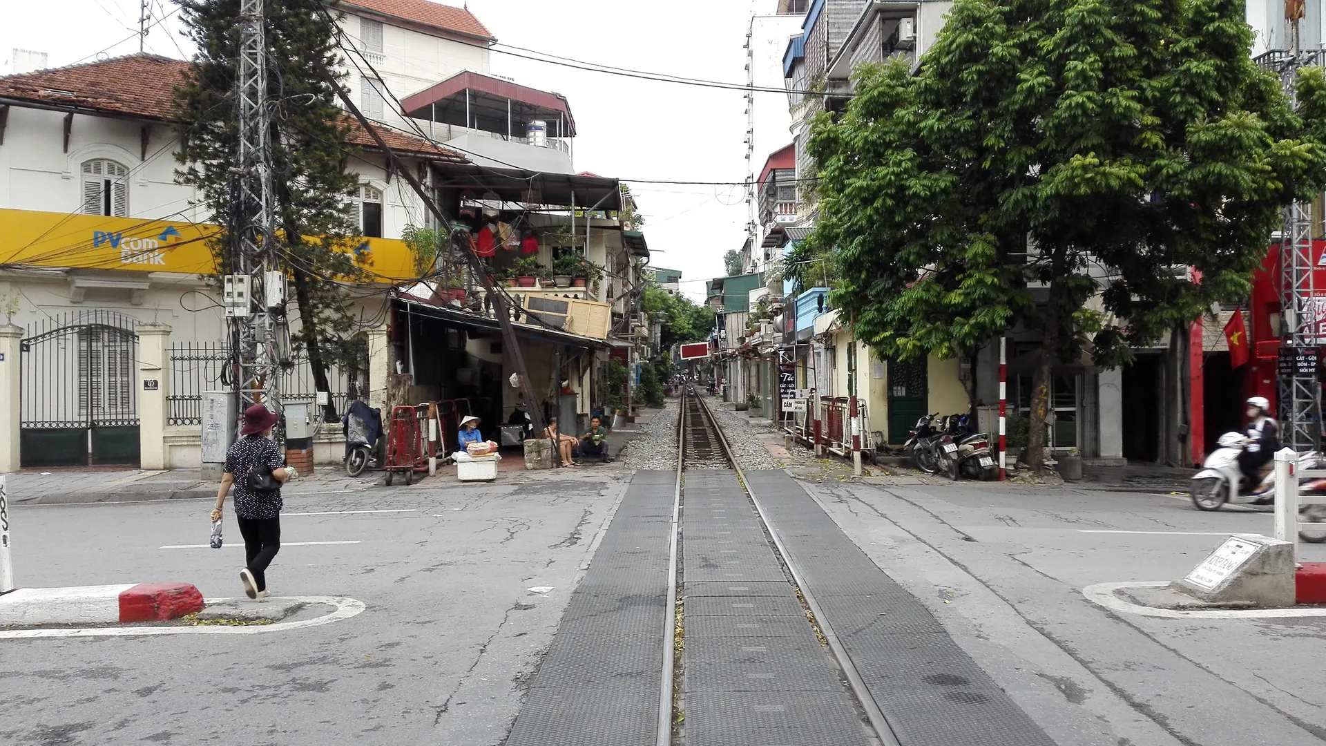 A railway track running straight through a street crossing in Hanoi, with shophouses, a bank and a scooter on either side of the rails