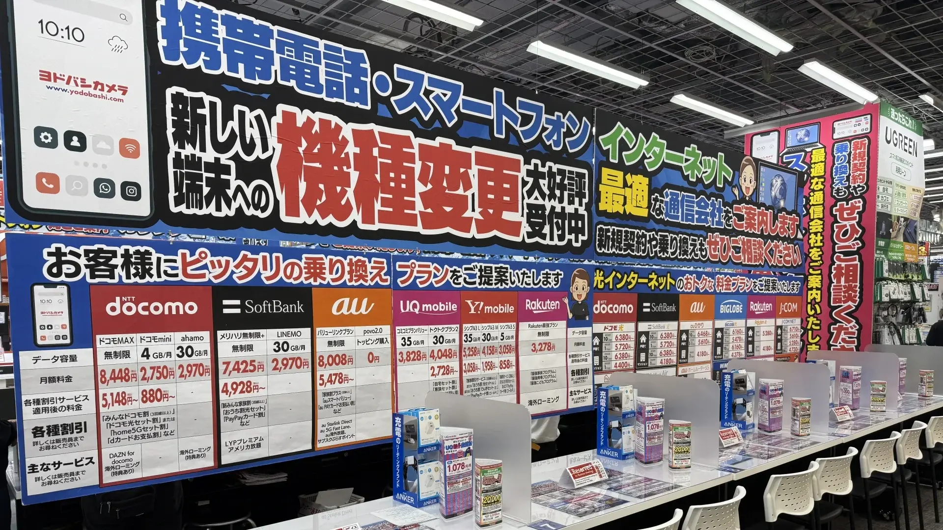 A mobile service counter in a Yodobashi Camera store, displaying large colorful banners and price lists for NTT Docomo, SoftBank, au and Rakuten Mobile