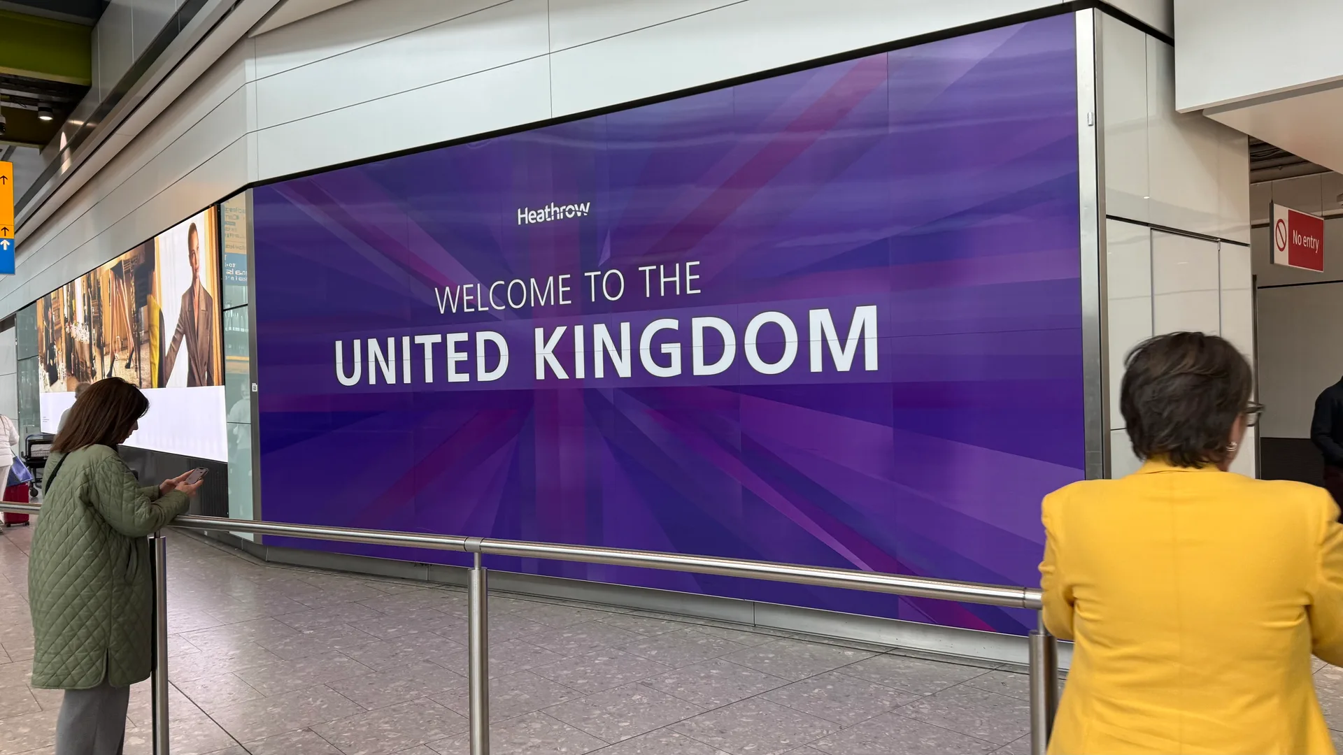 Large purple "Welcome to the United Kingdom" screen in the arrivals hall at London Heathrow, with an arriving traveler checking her phone