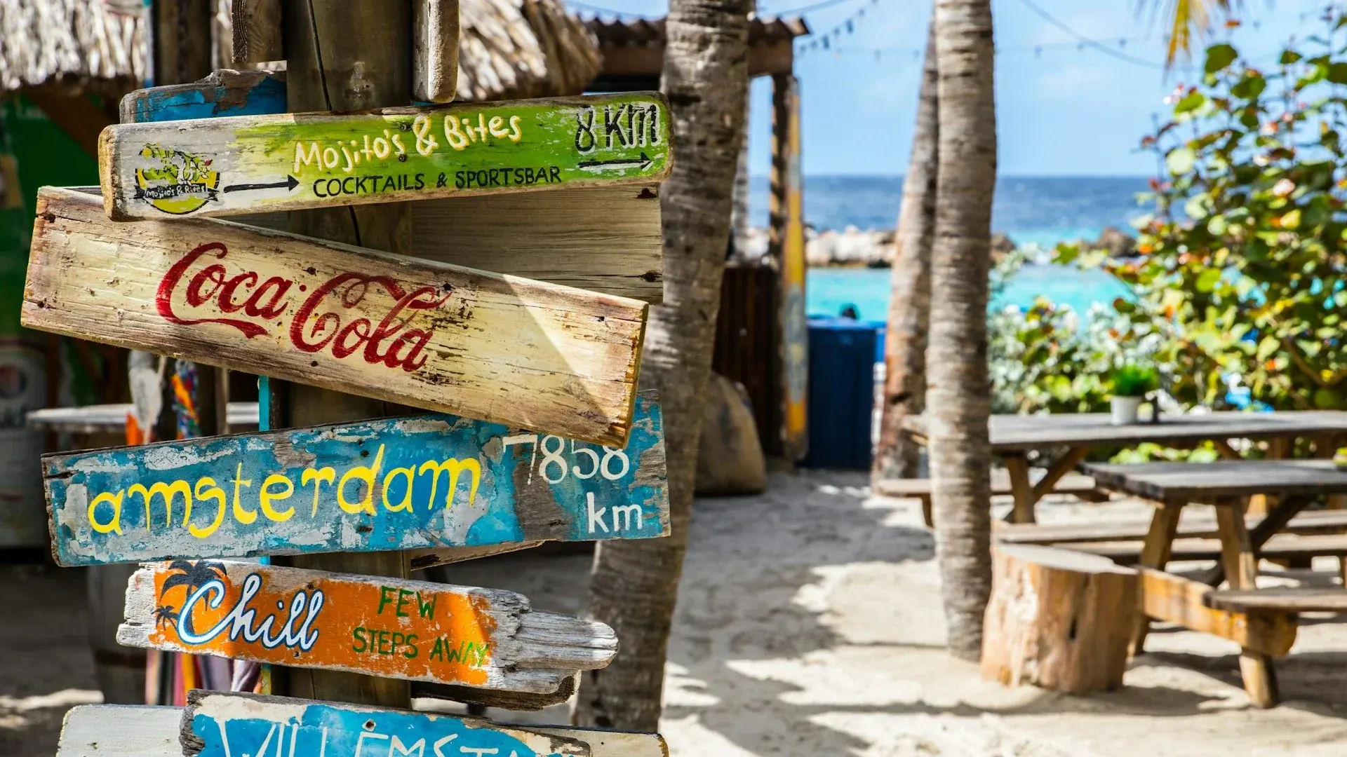 A colorful, rustic wooden signpost on a sunny beach in Curaçao, with painted signs pointing to destinations such as Amsterdam, Willemstad and local beach clubs.