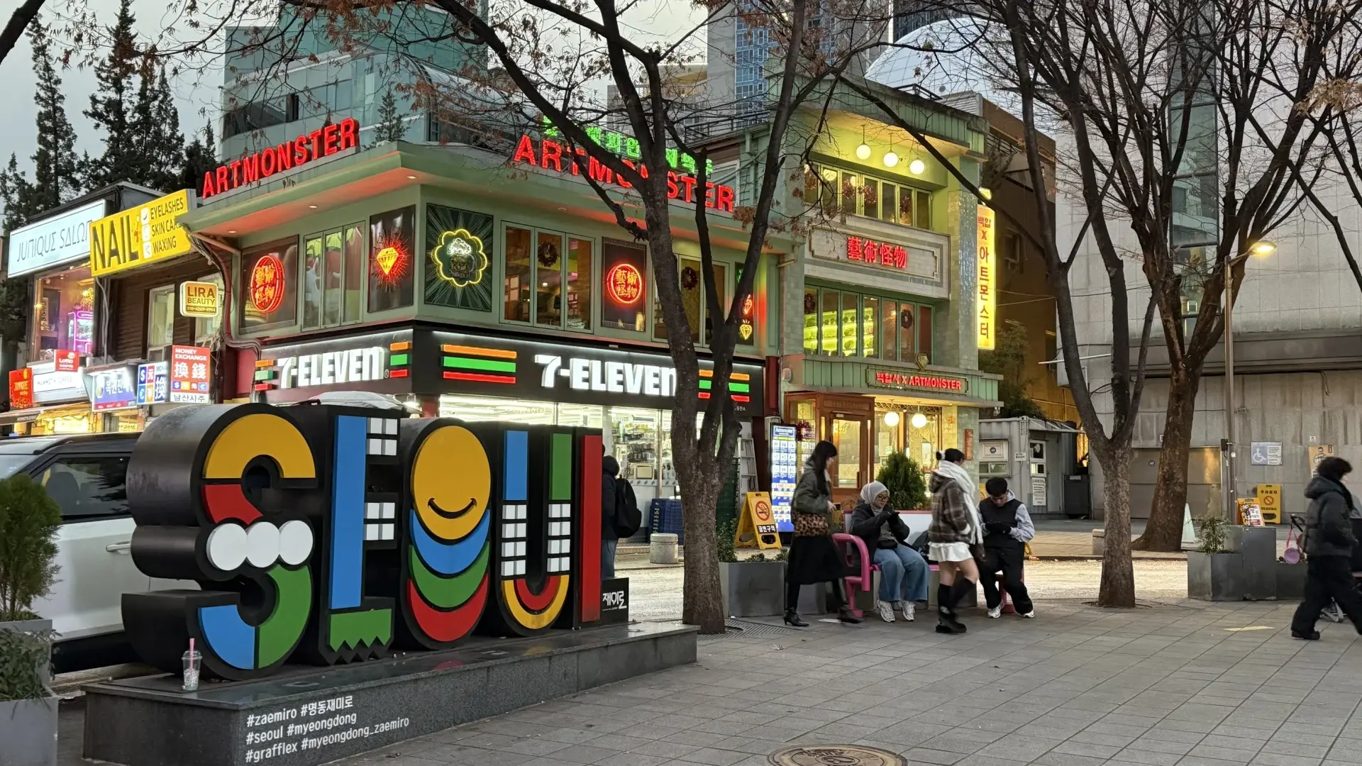 Colorful Seoul street sign in Myeongdong with neon shopfronts, a 7-Eleven convenience store and people sitting and walking in the evening